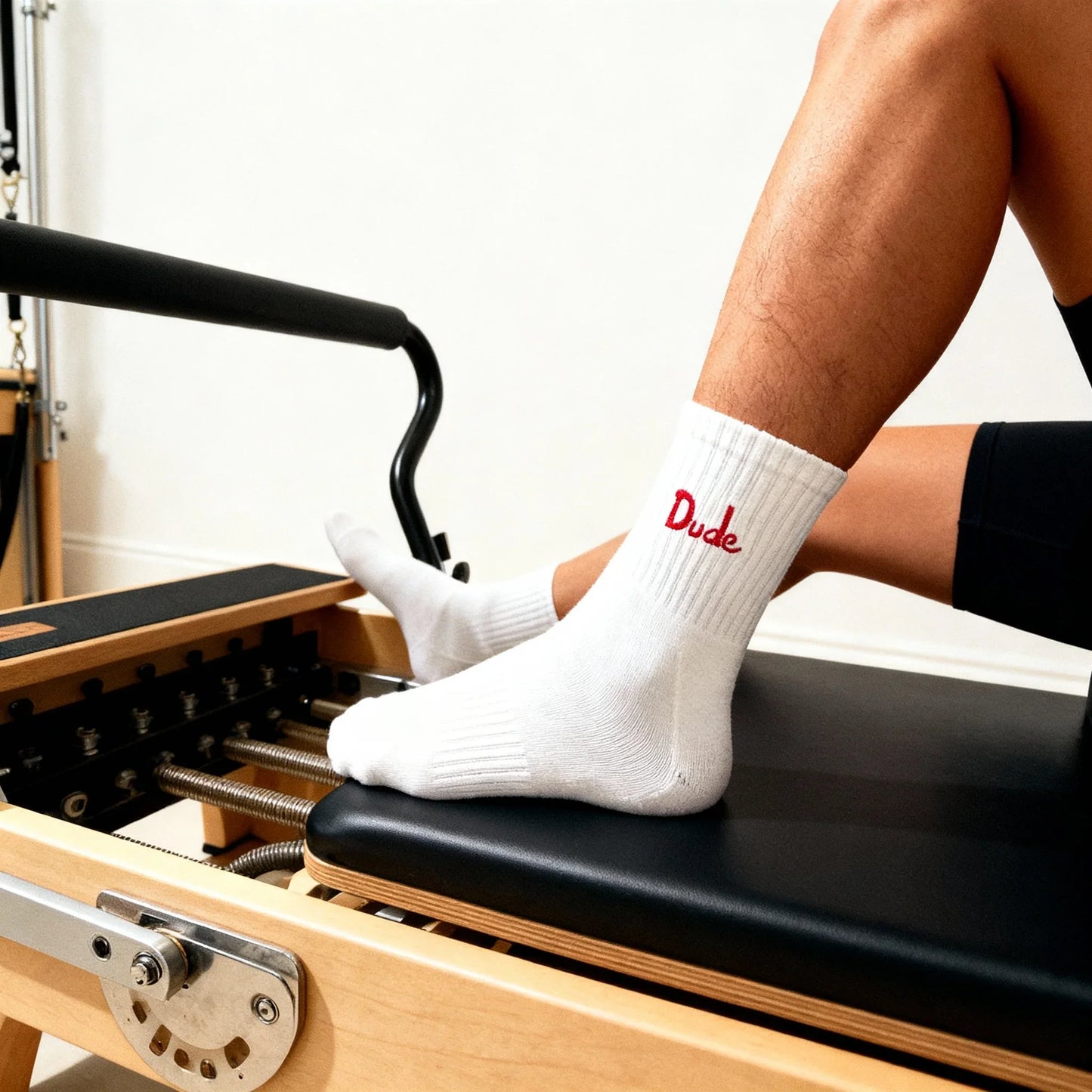 Close-up of a person wearing white socks with red "Dude" text, resting feet on Pilates reformer machine